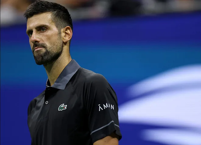 Close-up of Novak Djokovic in a black and grey Lacoste shirt during a night match, looking away from the camera with a blurred blue US Open backdrop behind him.