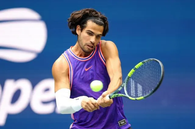 A tennis player hitting a two-handed backhand on a hard court at the US Open, wearing a purple and blue sleeveless shirt and a white sleeve on the right forearm.