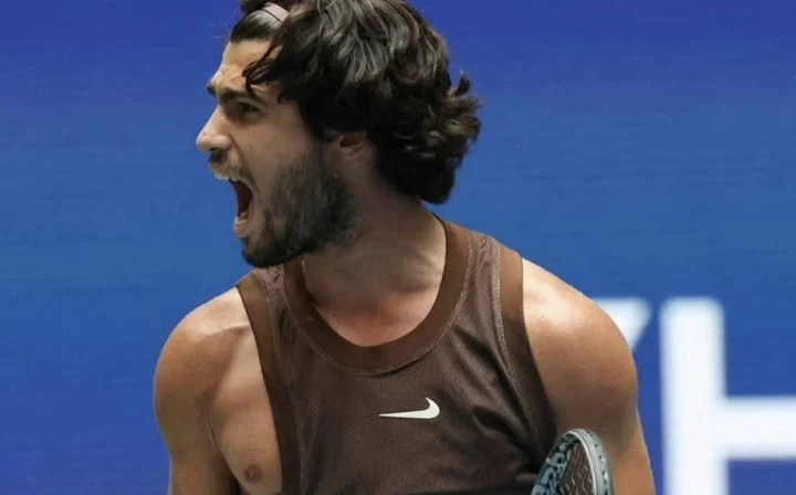 A tennis player celebrating in the brown sleeveless Nike top with a white swoosh on the chest, in front of the blue US Open backdrop.