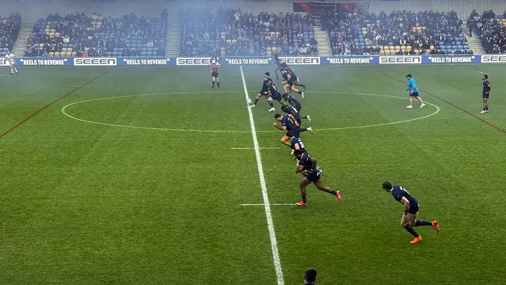 A rugby league kick-off in progress, the ball in flight above the halfway line with a chase line of players advancing