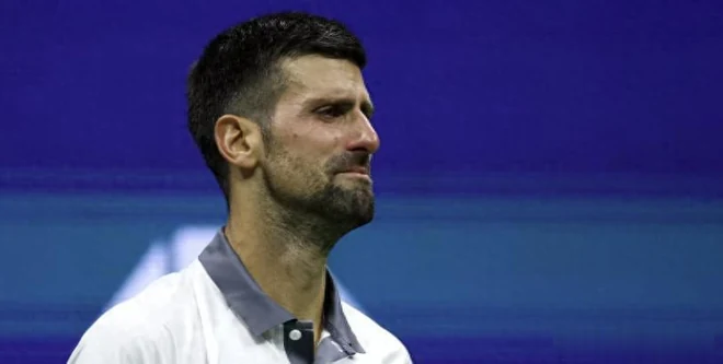 Novak Djokovic in a white and grey shirt with tears in his eyes, standing in front of the blue US Open backdrop.