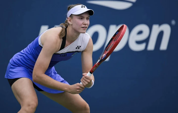 Elena Rybakina waiting to play a backhand in a blue dress and white visor, with the blue US Open logo wall behind her.